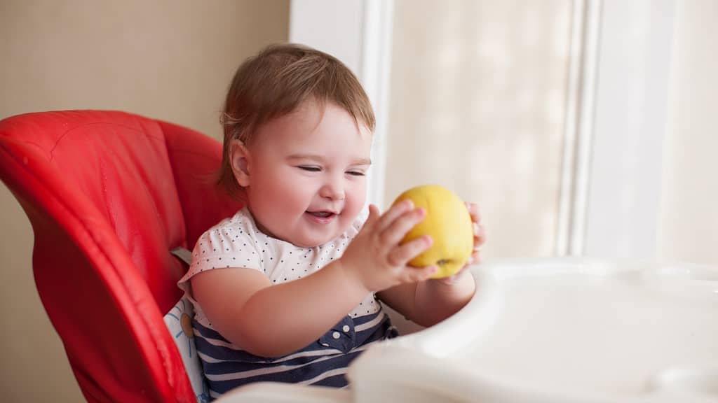 Small child sitting in a highchair with an apple in hand