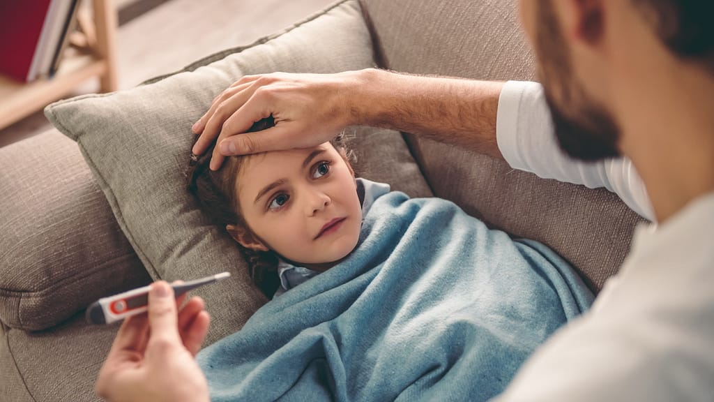 Sick child covered in blanket is lying on couch while father takes temperature