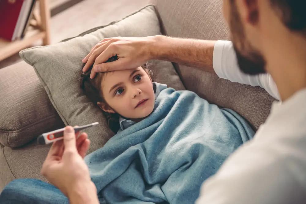 Sick child covered in blanket is lying on couch while father takes temperature